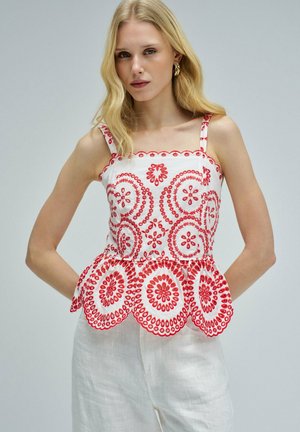 Femme blonde portant un top blanc sans manches avec une broderie circulaire rouge et un pantalon blanc, debout les mains dans le dos.