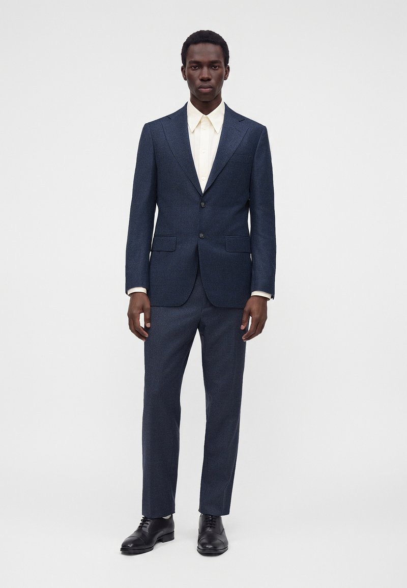 Homme portant un costume bleu foncé à deux boutons avec un pantalon assorti, associé à une chemise blanche à col et des chaussures habillées noires, debout immobile.