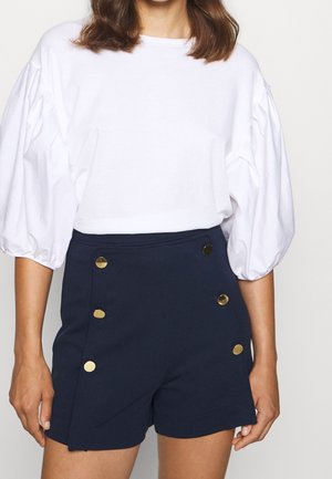 Femme portant un chemisier blanc à manches bouffantes et un short taille haute bleu marine avec des boutons dorés décoratifs sur le devant.