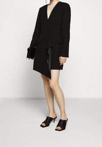 Robe blazer noire sur mesure avec un décolleté en V profond, un ourlet asymétrique et un détail à volants. Portée avec des sandales plates noires et un sac pochette noir.