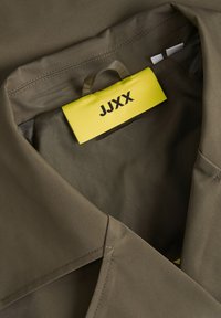Oliivivihreä kangastakki, jossa on lovettu kaulus. Kauluksen sisäpuolella on keltainen etiketti, jossa lukee "JJXX".