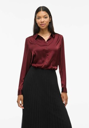 Blusa di satin bordeaux con colletto classico, maniche lunghe e chiusura con bottoni, abbinata a una gonna nera plissettata. Texture liscia e vestibilità su misura.