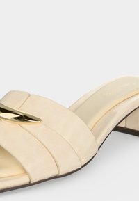 Sandale beige à semelle ouverte, avec de larges brides matelassées et un petit détail décoratif en métal doré sur le dessus, avec un talon bas à bloc.