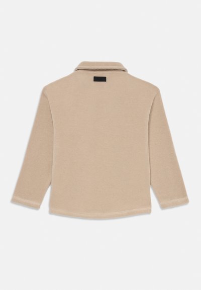 Beež pikkade varrukatega särk, mis on valmistatud pehmest kangast ja millel on krae, must silt ning kaarduv hem. Minimalistlik disain, ilma nähtavate mustriteta.