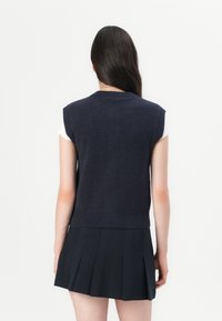 Maglione senza maniche blu navy con scollo rotondo e orlo a coste, indossato su una camicia bianca; presentato con una gonna nera plissettata.