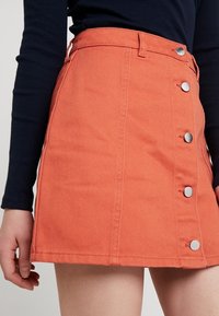 Mini falda de mezclilla en color naranja coral con una cintura ajustada, cinco botones a presión de plata y bolsillos laterales. Presenta un acabado liso y texturizado.