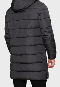 Manteau matelassé noir avec capuche, présentant un design matelassé, des manches longues et des détails zippés. Texture lisse et coupe mi-longue.