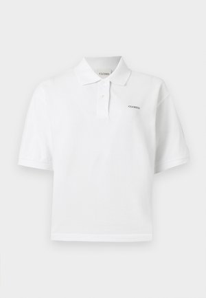 Άτομο που στέκεται φορώντας λευκό πόλο με κοντά μανίκια και φαρδιά μπλε παντελόνια με μαύρες κάθετες ρίγες, σε μονόχρωμο φόντο.