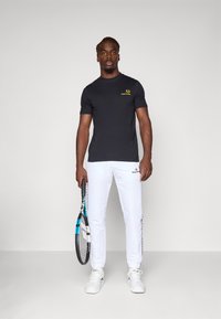 Sort kortærmet t-shirt med gult logo, hvide sweatpants med sidelogo, holder en tennisketcher, iført hvide sneakers.