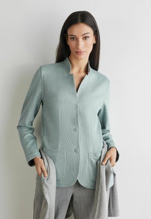 Frau mit langen dunklen Haaren trägt eine hellblaue Jacke mit Knöpfen über grauer Hose und hält einen passenden grauen Pullover in beiden Händen.