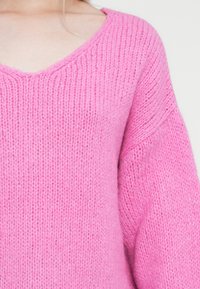 Maglione rosa lavorato a maglia con scollo a V, caratterizzato da un tessuto testurizzato e maniche ampie. Il filato appare morbido e peloso.