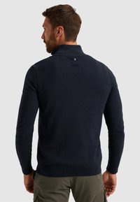 Marineblauwe gebreide trui met een hoge kraag en een accentdetail aan de achterkant. Getailleerde mouwen en tailleband, gemaakt van zacht materiaal.