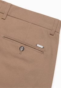 Zbliżenie na beżowe spodnie, ukazujące zapinającą się tylną kieszeń oraz małą metalową metkę z napisem "QUIET ESSENCE" po prawej stronie.