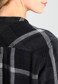 Persona bionda vista di spalle che indossa un giubbotto a trama nera e grigia, con il profilo del viso visibile di lato.