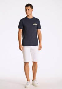 Tummansininen puuvillat-paita, jossa on graafinen taskuyksityiskohta, yhdistettynä valkoisiin shortseihin ja valkoisiin tennareihin. Paidassa on pyöreä kaula-aukko ja lyhyet hihat.