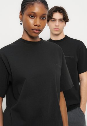 Twee jonge volwassenen dragen zwarte T-shirts met ronde hals en staan tegen een effen lichte achtergrond, de een voor de ander.