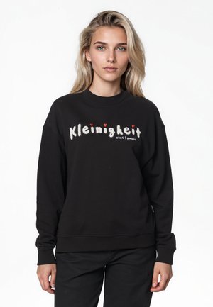 Ung kvinde med langt blond hår iført sort sweatshirt med teksten "Kleinigkeit avec l'amour" og små røde hjerter foran.