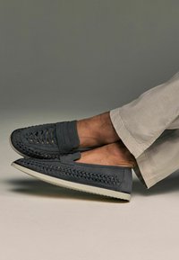 Los mocasines de cuero gris presentan un diseño trenzado, una banda ancha en la parte frontal y una suela de goma blanca, combinados con pantalones de color beige claro.
