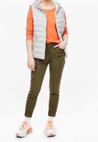 Lichtgrijze gewatteerde vest met capuchon over een feloranje langemouwen shirt, gecombineerd met olijfgroene slim-fit cargobroek en chunky witte sneakers.