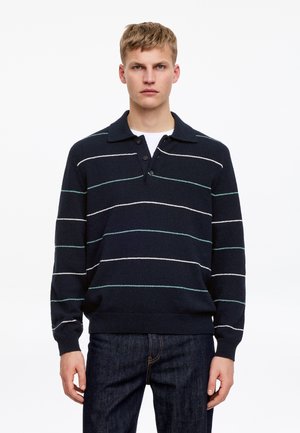 Ung mand iført en mørkeblå trøje med krave og hvide og turkise horisontale striber samt mørkeblå jeans, stående mod en hvid baggrund.