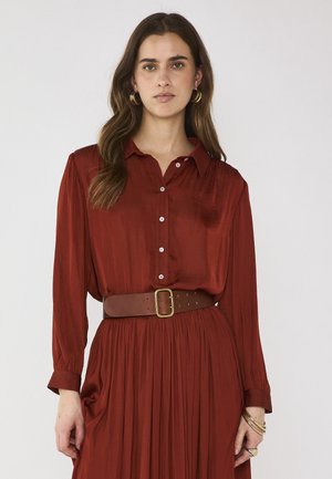 Donna con capelli lunghi e castani che indossa orecchini a cerchio dorati, un vestito a bottoni color ruggine con una cintura larga marrone e braccialetti dorati al polso.