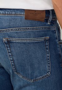 Jeans de mezclilla azul marino que cuentan con una etiqueta de cuero marrón con "Hackett London" en la parte trasera. Costuras visibles y un diseño estándar de bolsillo trasero.