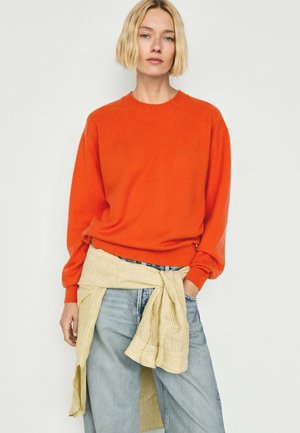 Kvinde med kort blond hår har en orange sweater, løse blå jeans og en lysegulstribet skjorte bundet om livet.