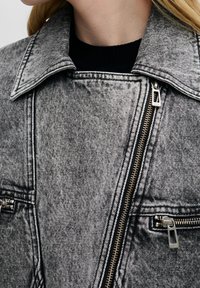 Grå denimjacka med en något sliten yta, som har en stor krage, silverfärgad dragkedjor och två fickor med dragkedja.