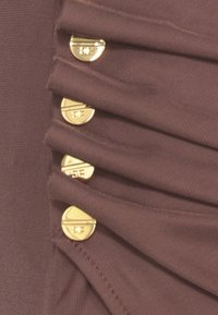 Tessuto marrone con pieghe, caratterizzato da cinque bottoni circolari in tonalità dorata lungo il lato, che conferiscono un aspetto strutturato e sartoriale.