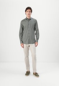 Grün gemusterte Button-Down-Bluse mit langen Ärmeln, cremefarbene Hose und grüne Slipper mit braunen Akzenten. Minimalistischer Hintergrund.