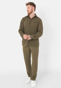 Olive green, long-sleeve button-up shirt with a collar, paired with matching joggers. Features logo detail on the shirt and pants. White sneakers.
