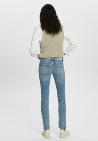 Stickad beige väst över vit skjorta, i kombination med smala ljusblå denimjeans och vita sneakers med strukturerade sulor.