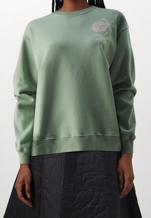 Vrouw draagt een saliegroene trui met een lichte bloemmotief op de borst en een zwarte doorgestikte rok.