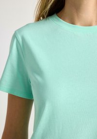 Närbild av en person som bär en enkel mintgrön t-shirt med korta ärmar och rund halsringning mot en ljus bakgrund.