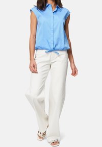 Blouse sans manches bleu clair avec un devant à boutons et un ourlet à cordon, associée à un pantalon large blanc et des sandales. Texture de tissu lisse.