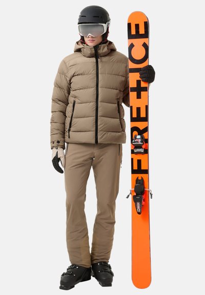 Ski outfit featuring a tan puffer jacket, beige pants, black gloves, and ski boots. Holding an orange ski with black lettering.