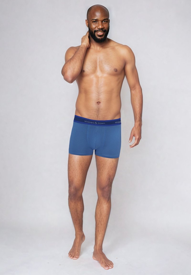 Boxer da uomo blu in cotone con un elastico scuro a contrasto che presenta il nome del marchio. Design semplice e texture morbida. Modello in piedi in modo casual.