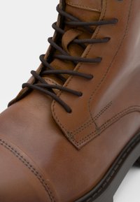 Bota de cuero marrón con un acabado suave, puntera redonda y cordones marrones. Presenta costuras detalladas y una suela resistente.