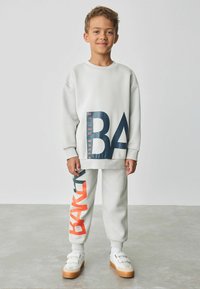 Hellgrauer Sweatshirt und Jogginghosen mit großen navyblauen und orangenen Textgrafiken. Glatter Stoff mit gerippten Säumen und Bund. Weiße Sneakers.