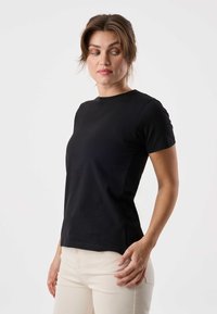Vrouw draagt een strakzittend zwart shirt met korte mouwen en lichtbeige broek, kijkt zijwaarts tegen een effen witte achtergrond.