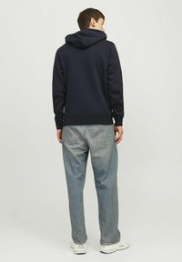 Hoodie navy con cappuccio regolabile, polsini e orlo a costine, abbinato a jeans larghi azzurri e sneakers bianche. Sfondo semplice.