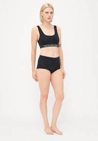 Sort sports-bh og matchende boyshorts med en glat tekstur. Bh'en har et reflekterende logo bånd. Modellen står barfodet og vender fremad.