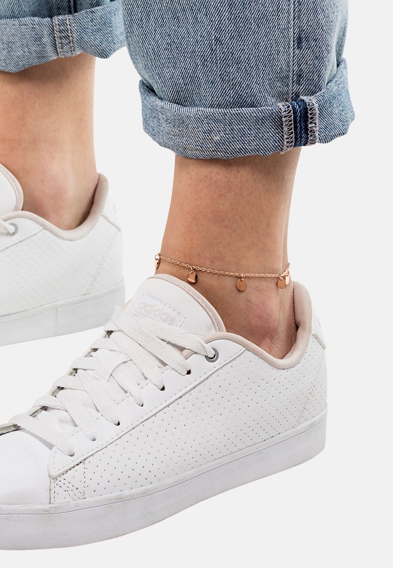 Cheville avec un délicat bracelet chaîne en or avec de petits charms ronds, portant un jean bleu clair retroussé et des baskets blanches Adidas.