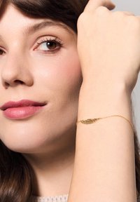 Złoty bransoletka z piórami o skomplikowanych detalach, leżąca na jasnej karnacji. Design łańcuszka jest delikatny, z małym zapięciem.