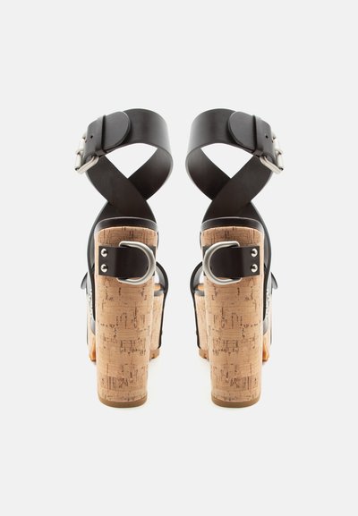 Πέδιλα με ψηλό τακούνι από φελλό, μαύρα δερμάτινα λουράκια που διασταυρώνονται στον αστράγαλο και ασημένιες αγκράφες, εμφανιζόμενα από πίσω.