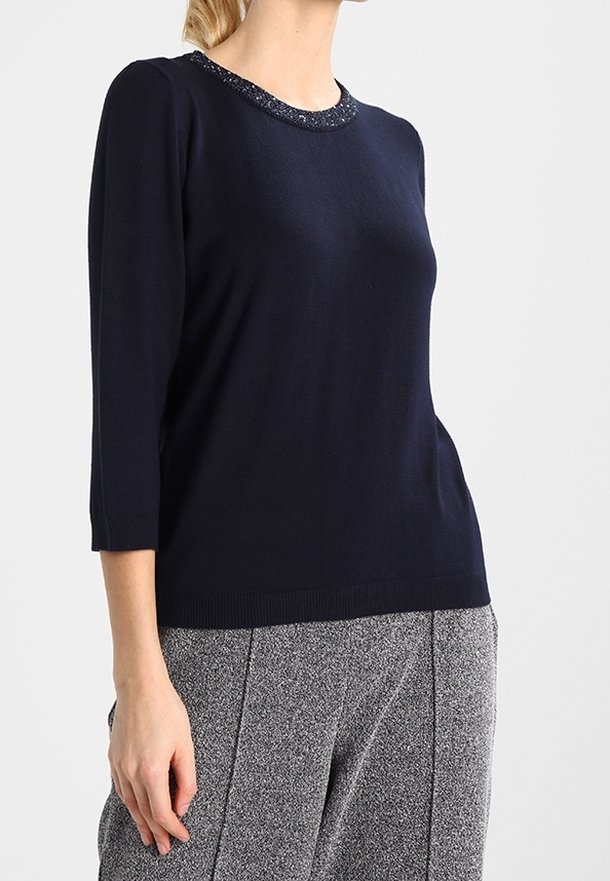 Femme portant un haut à manches longues bleu marine avec un col orné de perles et un pantalon gris texturé, debout devant un fond uni.