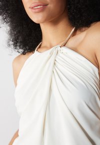 Balts halter tops no teksturēta auduma, ar drapētiem elementiem un zelta metāla detaļu pie kakla daļas.