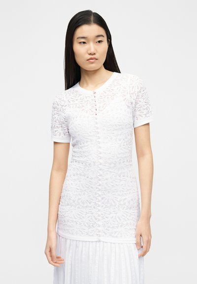 Mladá žena v bílé krátkém krajkovém topu s látkou potaženými knoflíky vpředu a bílé plisované sukni.