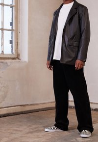 Svart läderblazer, vit skjorta, svarta kordfläskbyxor och svart-vita rutiga slip-on sneakers, mot en texturerad vägg.