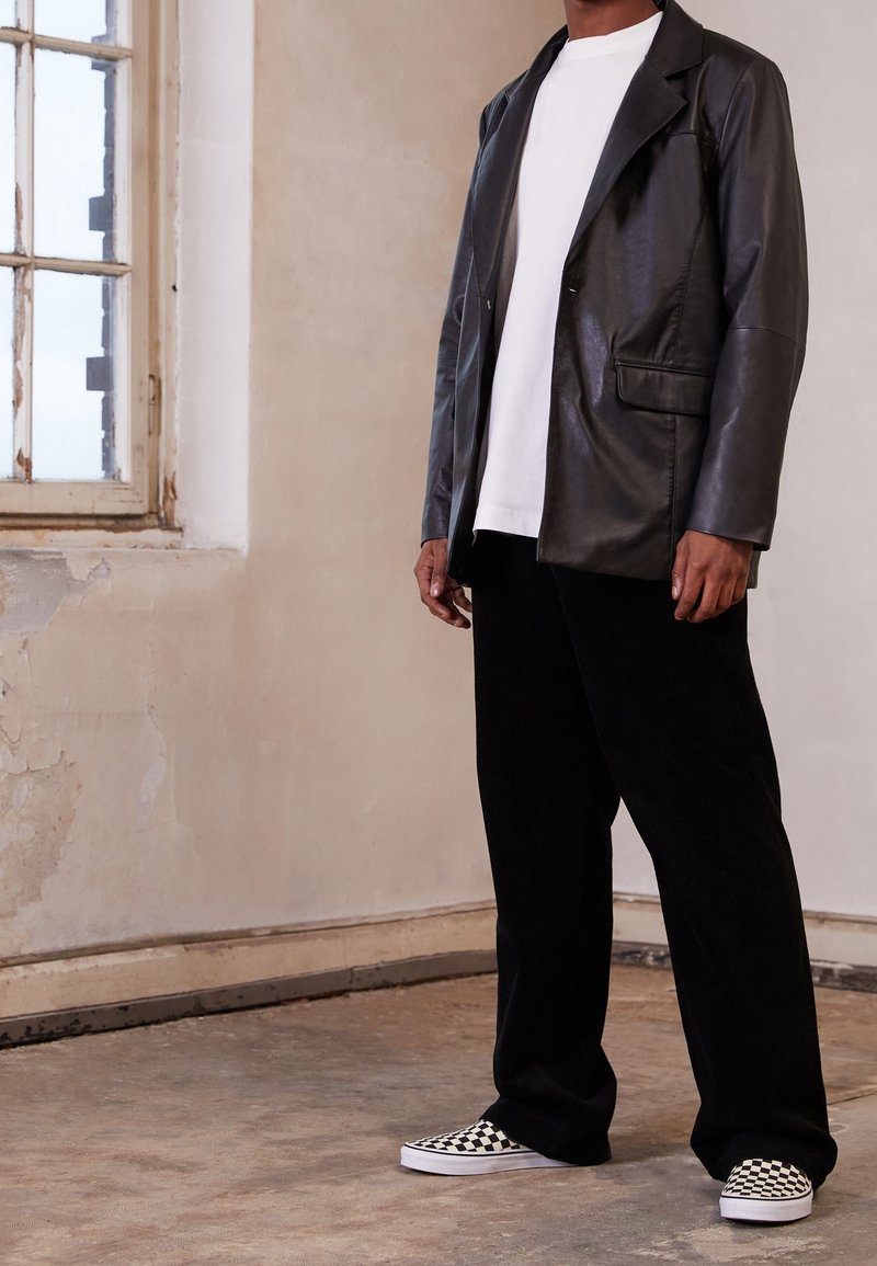 Svart läderblazer, vit skjorta, svarta kordfläskbyxor och svart-vita rutiga slip-on sneakers, mot en texturerad vägg.